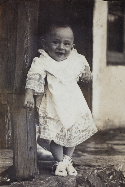 Sonny Hutchinson, wearing a lace baby gown, being lifted to stand on a summer house verandah, Moganshan