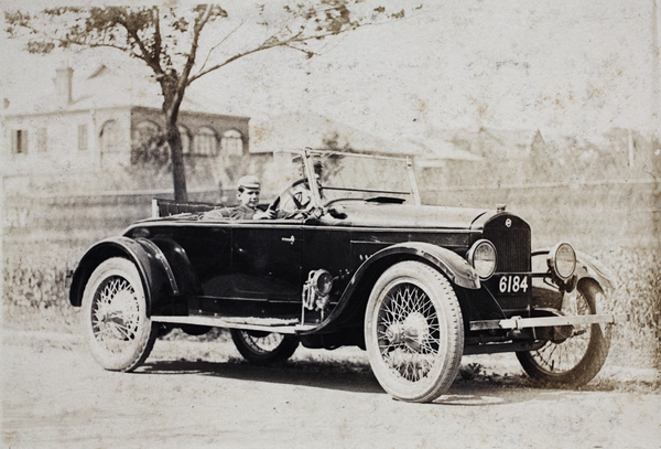 Baba Hansen posing behind the wheel of an automobile, with a uniformed driver in the front passenger seat, Dalney Road, Hongkou, Shanghai