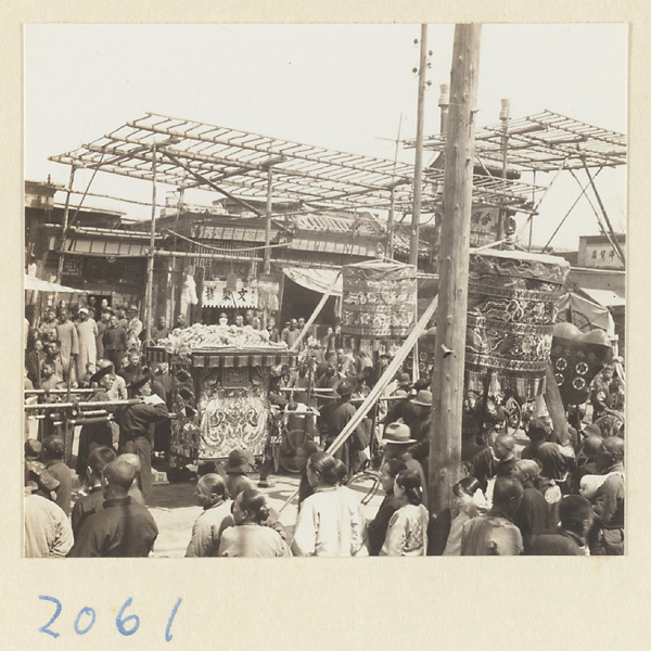 Members of a wedding procession carrying sedan chair, umbrellas, and fan-shaped screen past shops