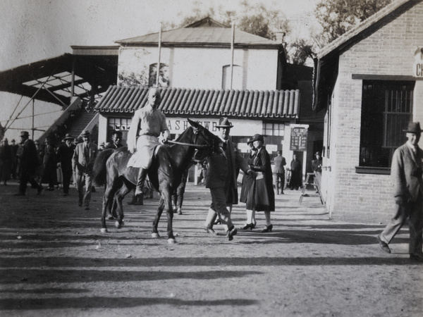 A man leading a horse past the 'CASH SWEEP' betting booth, Peking Races, Beijing