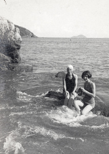 Maggie Henderson and Gladys Hutchinson, wearing bathing costumes and sitting on a rock in a bay, Hong Kong 