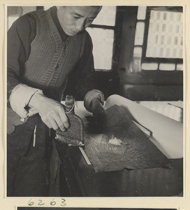 Interior of a scroll-mounting shop showing a man drawing paste from a piece of scrap paper to paste the edge of a scroll