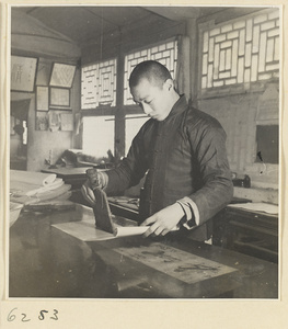 Interior of a scroll-mounting shop showing a man adhering a backing to a scroll by softly tapping with a stff brush called a zong pi shua
