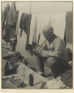 Clothes hawker sitting with his wares and smoking a pipe