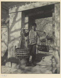 Rag and bone dealer with baskets suspended from a shoulder pole standing in a doorway with auspicious couplets