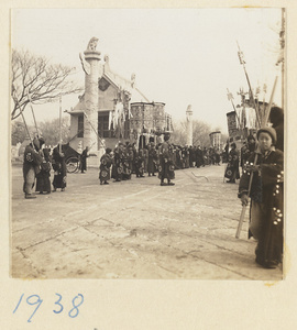 Members of a funeral procession carrying umbrellas, staffs, and banners past building with hua biao