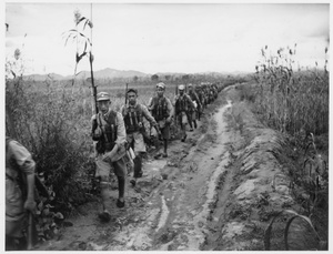 Soldiers (in vanguard) walking  along a track beside fields with crops, 1938