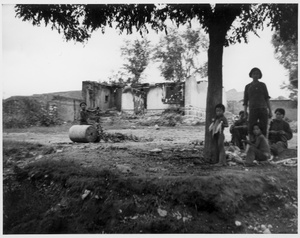 Burnt down houses, with a women and children outside, 1938