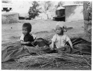 Yinhua, the baby son of Guo Qinglan and Dr Dwarkanath S. Kotnis, sitting on a  sheet on top of some hay, with another baby, Jinchaji